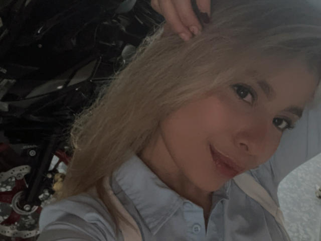 AmaraRicci, avec ses cheveux blonds, souriante, portant une chemise bleue, tenant ses cheveux d'une main, des pièces de moto visibles en arrière-plan. Ses fesses ne sont pas visibles sur l'image..