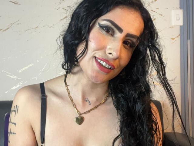 DemiMistral with long black wavy hair, wearing a black strap top, gold necklace, and red lipstick, showing off her tits and tattoos, smiling at the camera.