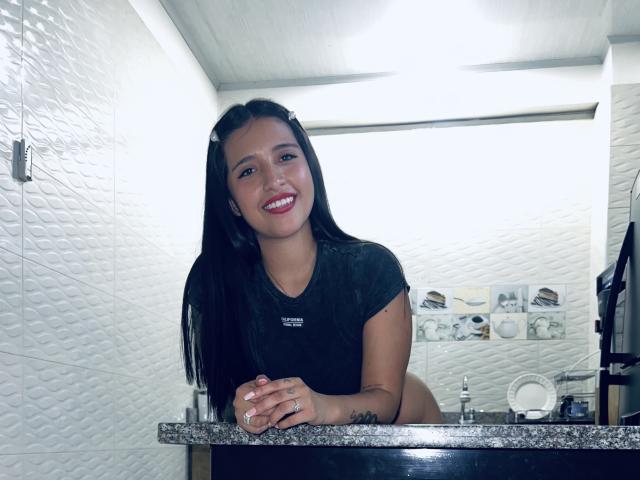Sophie, avec de longs cheveux noirs, souriante, portant un t-shirt noir, debout derrière un plan de travail en granit, les mains posées dessus, tatouage visible sur l'avant-bras droit, murs en carreaux blancs..