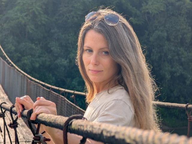 AuroraStar with long brown hair, sunglasses on head, white shirt, holding a rope on a bridge, looking over her shoulder with a slight smile. Forest in background.