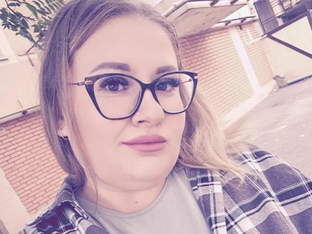 RadaRori has big tits, wears black-framed glasses, and has light brown hair in a plaid shirt over a gray tee. She's outside with a brick wall in the background.