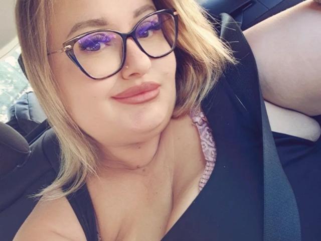 RadaRori has purple eyeliner, black glasses, and blonde hair, wearing a black top with a seatbelt. Her tits are visible, and she's sitting in a car.
