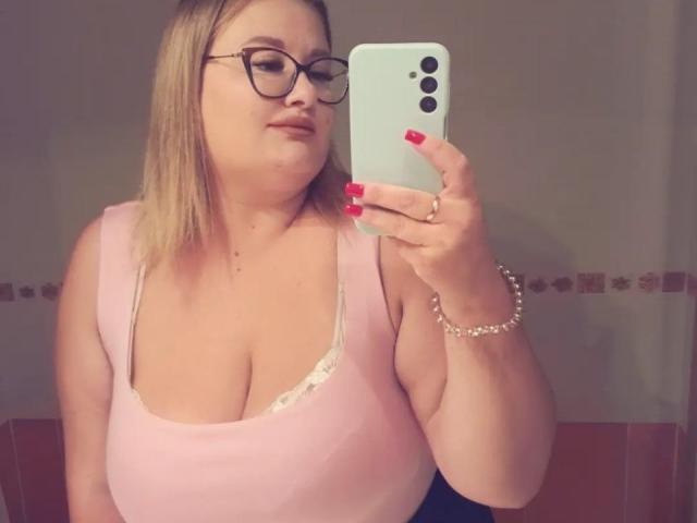 RadaRori with glasses, blonde hair, pink tank top showing big tits, white lace, red nails, pearl bracelet, taking selfie in mirror with iPhone.
