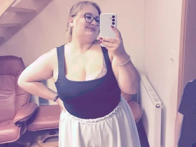 RadaRori with glasses, black tank top showing large boobs, lace bra peeking, white skirt, smiling, holding phone, standing in room, pink leather chair in background.