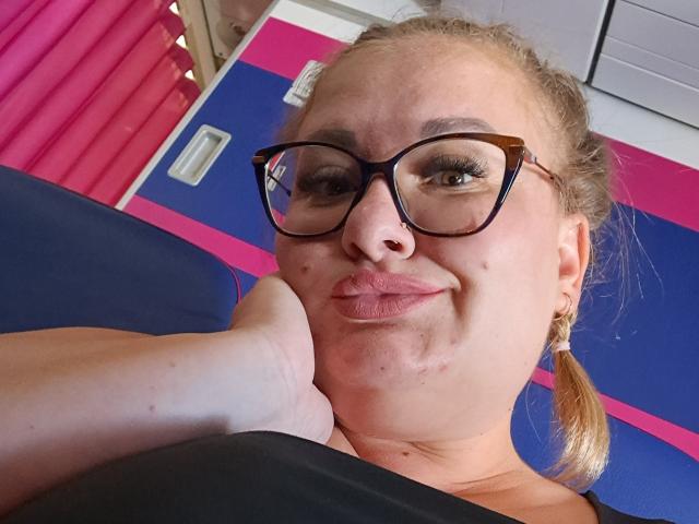 RadaRori with glasses, puffy lips, blonde pigtails, holding her own cheek, blue and pink wall in background, black top, slightly chubby, smiling, upper body shot.