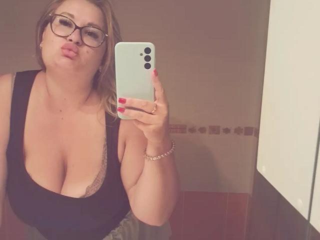 RadaRori with glasses, pouty lips, and red nail polish, holding a phone, wearing a black top with deep cleavage showing large tits, in a mirror selfie.