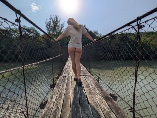 AuroraStar standing on a wooden, netted bridge, facing away, wearing a white t-shirt and black thong, showing her ass, with sunlit water and trees in the background.