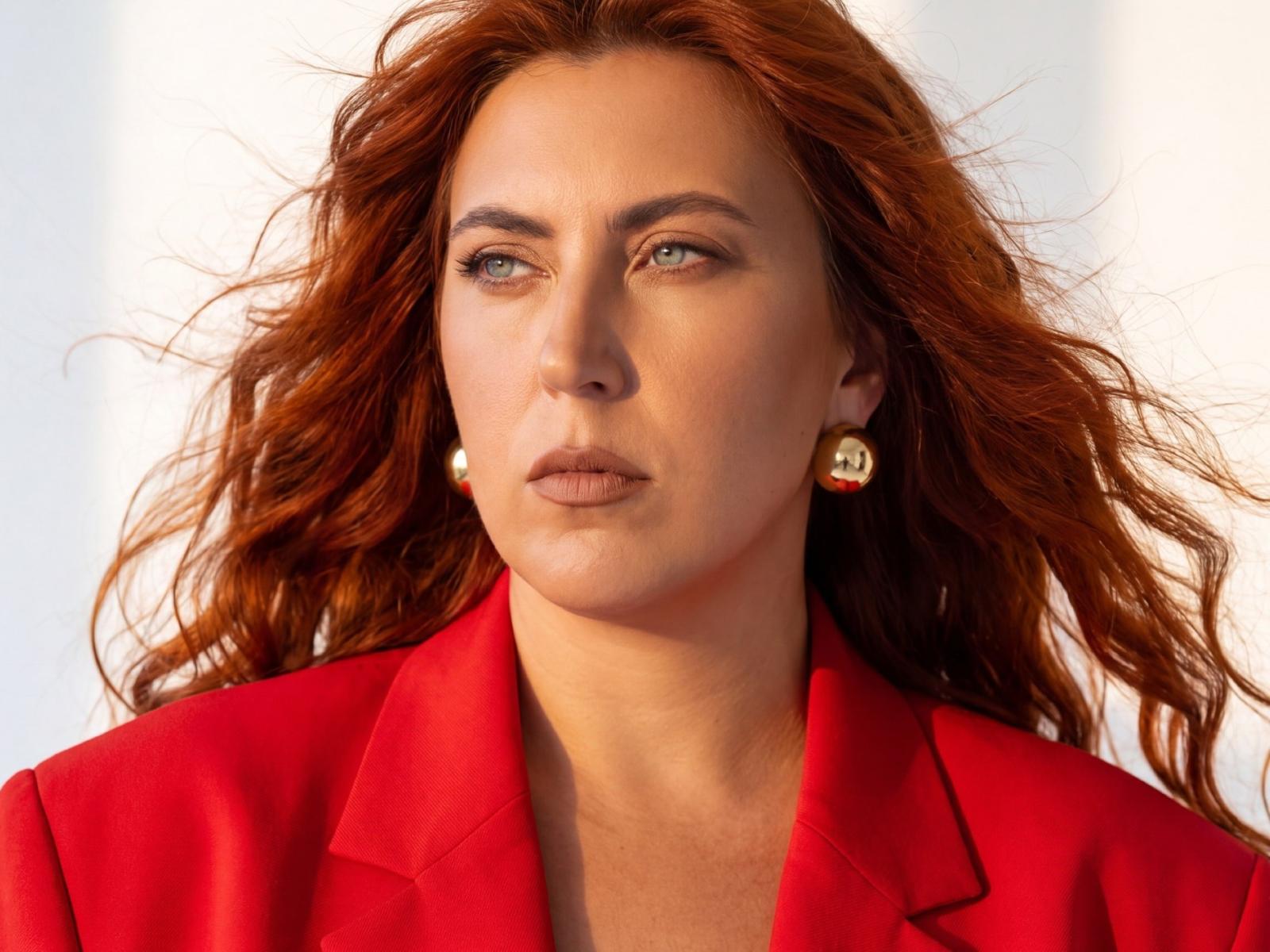 IsabellaFabbinin has red wavy hair, blue eyes, and a serious expression, wearing a red blazer with gold earrings, looking slightly to the side.