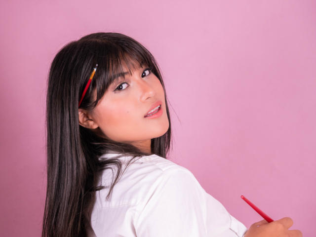 AshlyLee con lunghi capelli neri, camicia bianca, che tiene una matita rossa, guarda oltre la spalla, sfondo rosa, sorriso dolce, trucco leggero, naso piccolo, carina, sexy..