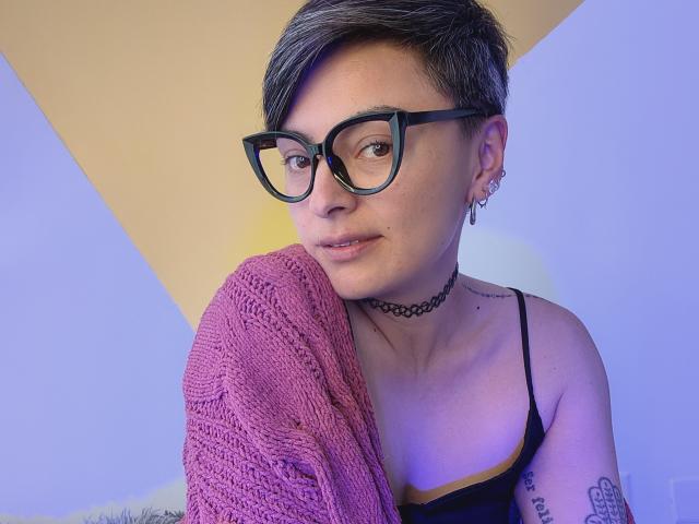 KatParadise mit kurzen, braunen Haaren, schwarzen Brillen, Choker, Tattoos, hält einen rosa Strickpullover, ein schwarzes Top mit Trägern, blickt in die Kamera. Sichtbar kleine Brüste, Ohrringe, ein leichtes Lächeln..