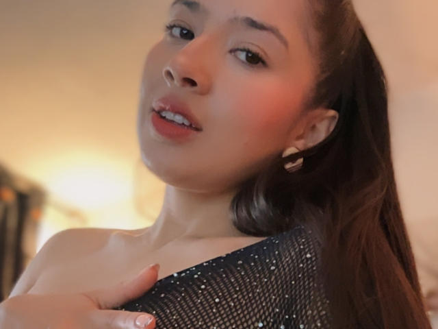 EmmaLombardi with long brown hair, looking at camera, holding black mesh top, showing shoulder and nipple, soft smile, brown eyes, subtle makeup, gold earrings, warm lighting.