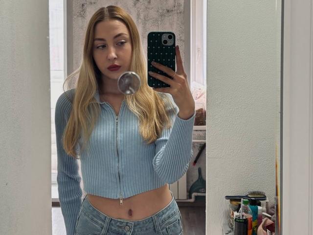 NettieHantz with blonde hair, blue ribbed crop top, and blue jeans, blowing bubble gum, holding phone with green case, mirror selfie, visible belly button, neutral expression.