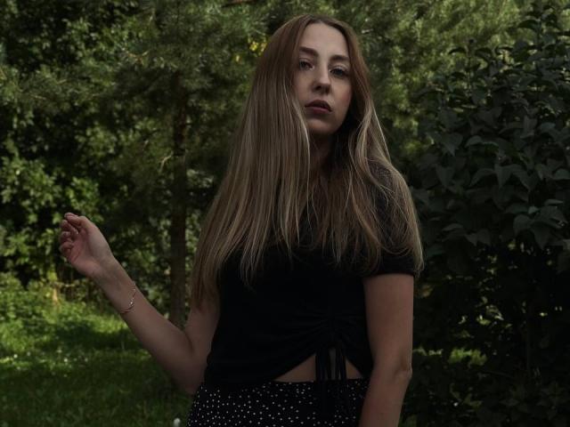 NettieHantz with long brown hair, black shirt, black polka dot skirt, standing in greenery, right arm raised, looking slightly to the side, dark lipstick, subtle expression.