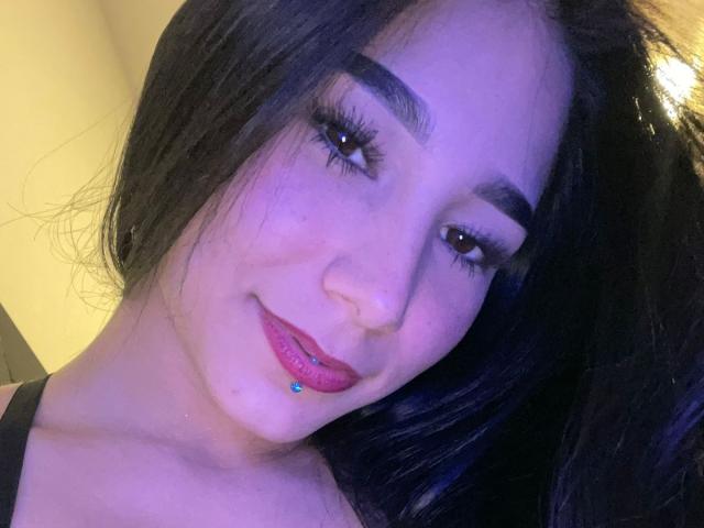 VioletaVelvet has dark hair, thick eyebrows, long eyelashes, and a slight smile with pink lipstick and a blue lip piercing, looking at the camera. She's wearing a black top.