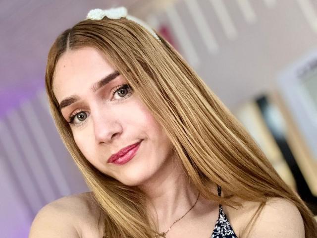 AndyDupont has long, straight blonde hair with a white hairband, brown eyes, and pink lipstick. She's wearing a black-and-white patterned top, showing her bare shoulders.