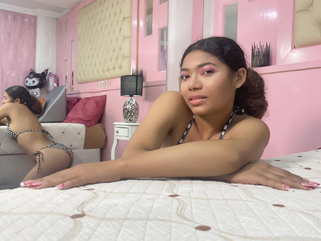 Sheillany, avec sa peau bronzée, son fard à paupières rose et ses cheveux noirs, portant un bikini rayé noir et blanc, est appuyée contre un lit, montrant ses seins et ses fesses, avec des ongles roses, dans une pièce rose..