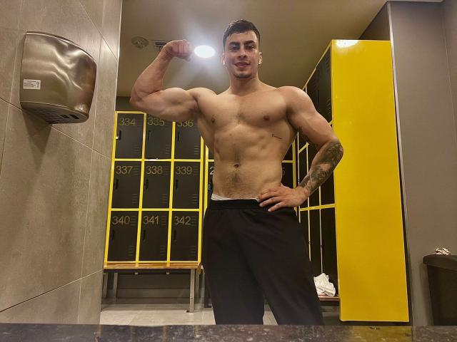 ThiagoMeneses, muscular and shirtless, flexes his right bicep, showing off his tattoos and black pants, standing in a locker room with yellow lockers in the background.