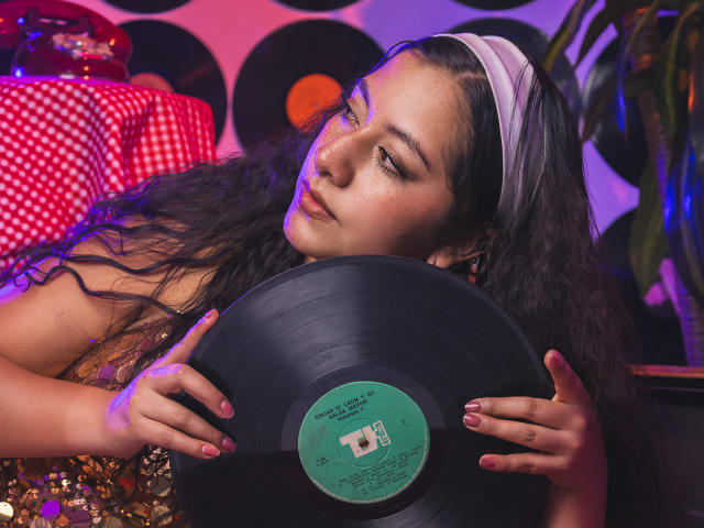 PaulaLoughty with curly black hair, wearing a white headband, holding a black vinyl record against her face, in a colorful, retro-themed setting, looking slightly bored.