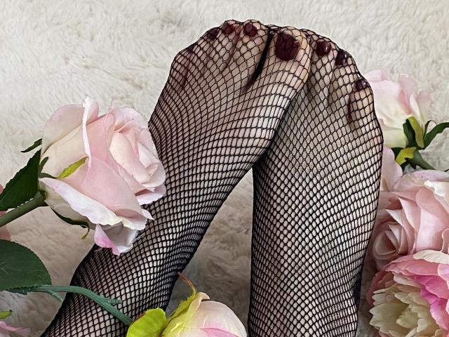 MiraRosse's feet in black fishnet stockings, with dark red toenails, crossed at the toes, surrounded by pink and white roses.
