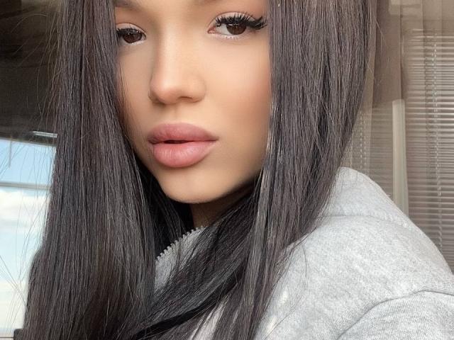 StevieMilman has long, straight, dark brown hair, full pink lips, and brown eyes with long lashes. She's wearing a gray hoodie, looking directly at the camera.