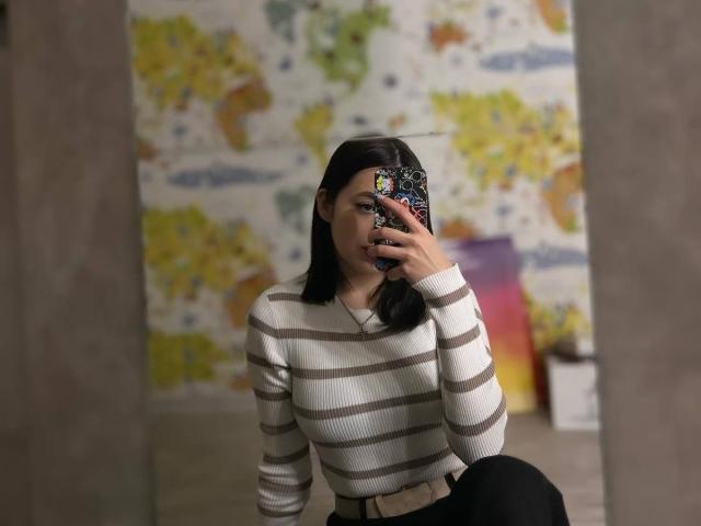 StevieMilman with black hair, wearing a striped white and brown sweater, holding a phone with a colorful case, sitting with one knee up, in front of a mirror.