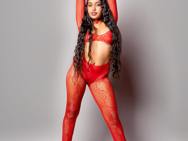 JeimyCampos with long curly black hair, wearing a red lace bra, red high-waist pant, and red fishnet arms, standing with arms raised, showing her tits and red choker.