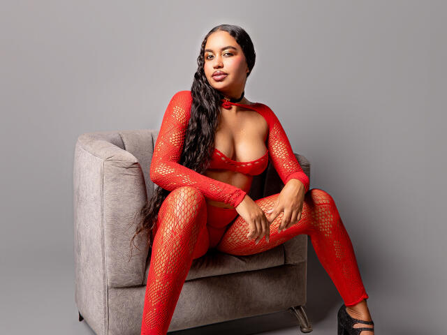 JeimyCampos in red fishnet outfit, sitting on gray chair, legs spread, showing her tits, with long black hair, black heels, and a confident expression.