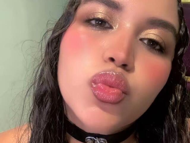 JeimyCampos with glossy pink lips, pouty face, and wet dark hair, wearing a black choker, has shiny gold eyeshadow and rosy cheeks, looking seductively into the camera.