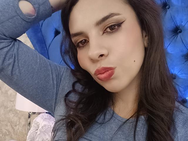VioletaStone, avec ses lèvres charnues, ses cheveux ondulés foncés et son eye-liner ailé, portant une chemise bleue avec des manches déchirées, une main dans les cheveux, un fond bleu texturé, une peau pâle, de jolies taches de rousseur..