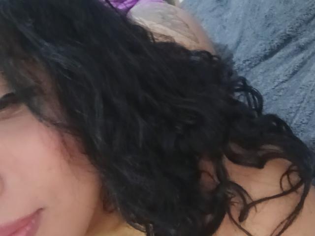 MariaCatalina with curly black hair, light skin, and a partial view of her face, lying down, showing a bit of her bare shoulder and purple bra.