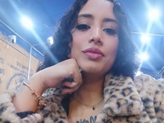 MariaCatalina with curly black hair, wearing a leopard fur coat, pink lipstick, and gold bracelet, resting her chin on her hand, looking at the camera.