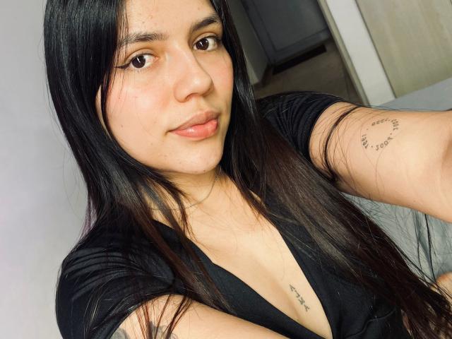 SophiaMarx with long black hair, brown eyes, and a small tattoo on her right arm, wearing a black V-neck shirt, showing her tits and a chest tattoo.