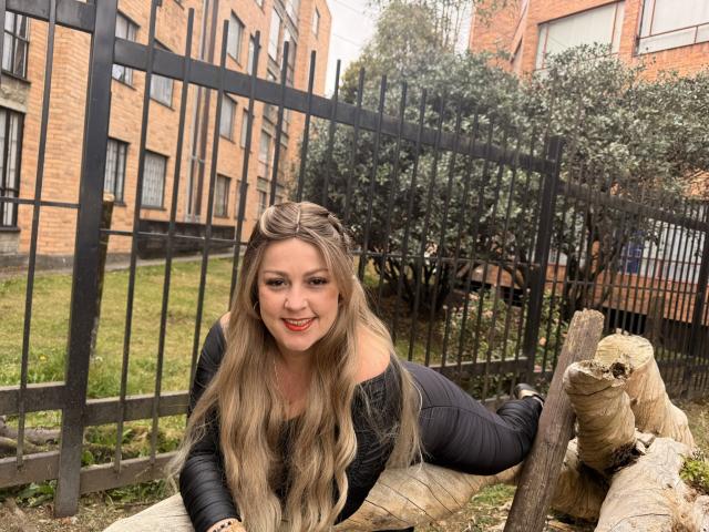 SamanthaGold with long blonde hair, red lipstick, black off-shoulder top, and black pants, laying on logs, smiling, hands on log, black metal fence, brick building in background.