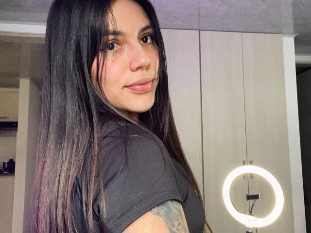 SophiaMarx with long black hair, brown eyes, and pink lips, wearing a black shirt, showing a tattoo on her arm, standing in front of a ring light.
