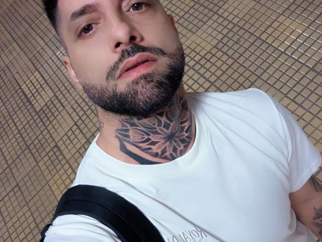 TommyDum con barba, tatuaggi sul collo, maglietta bianca, zaino nero, che guarda la fotocamera, capelli scuri corti, occhi marroni, bocca leggermente aperta, parete con piastrelle sullo sfondo..