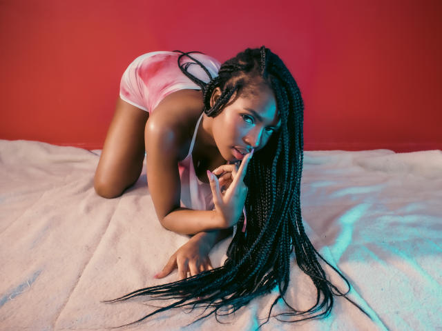 EllysRose è a quattro zampe, indossa pantaloncini rosa, ha lunghe trecce, succhia il dito, guarda la fotocamera, è sdraiata su una coperta bianca, in una stanza illuminata di rosso..