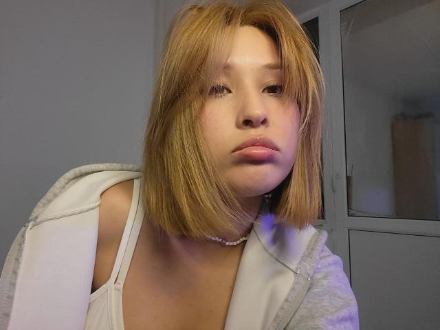 Chantell Houlihan, avec des cheveux blonds mi-longs, des lèvres pulpeuses, portant un débardeur blanc et un sweat à capuche gris, un collier visible, regardant légèrement sur le côté, expression neutre..