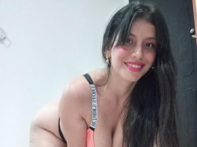 Madisson Fontaine, avec ses longs cheveux noirs, ses joues roses et son rouge à lèvres rouge, souriante, portant un soutien-gorge noir et rose avec l'inscription "LITTLE MISS" sur le bandeau, montrant ses grandes seins..