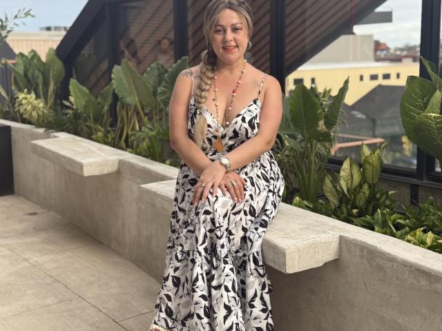 SamanthaGold with blonde braid, wearing a black and white floral dress, sitting on a concrete planter, hands on her knees, smiling, her tits partially visible, necklace on.