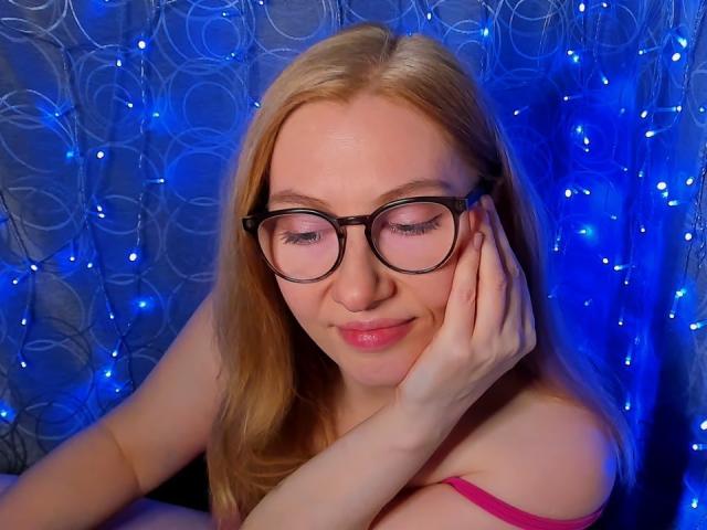 PaulinaSunny with glasses, blonde hair, pink lips, smiling softly, hand on cheek, wearing pink strap, blue fairy lights, and white patterned curtain in the background.