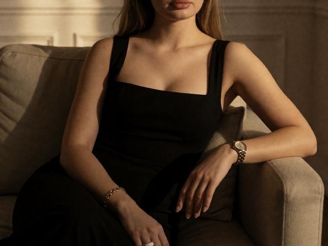 Destinis is sitting in a chair wearing a black dress with square neck, showing her tits, and a silver watch on her right wrist. Her hand rests on her thigh.