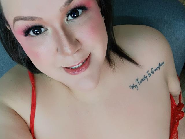 GiiannaMorrys com sombra rosa, delineador preto, batom vermelho e uma tatuagem no ombro direito. Ela está usando um sutiã de renda vermelho, mostrando seus seios. Ela está sorrindo para a câmera..