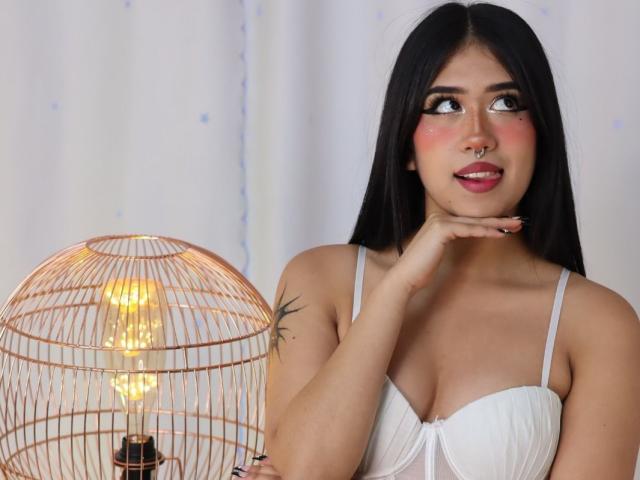 HannaThorne with black hair, white bra, and nose ring, holding a wicker birdcage with a glowing light bulb, looking to the side with a slight smile.