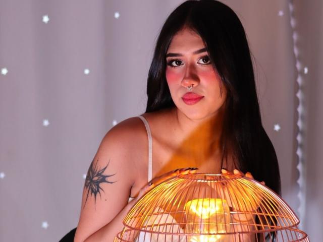 Hanna Thorne, com cabelos pretos longos, piercing no nariz, blush rosa, segurando um abajur de gaiola de arame amarela, tatuagem no ombro direito, vestindo um vestido branco com alças, olhando para a câmera..