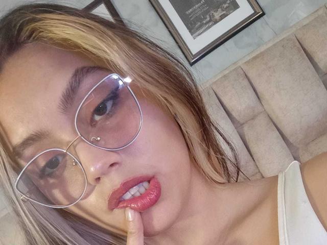 RubyLane with blonde highlights, wearing glasses, has pink lipstick, a nose ring, and is biting her finger, showing off her tits in a white top.