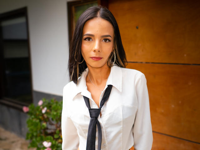 EvelinLuna with straight dark brown hair, wearing a white button-up shirt and black tie, standing in front of a wooden door, has small tits and wears large gold earrings.