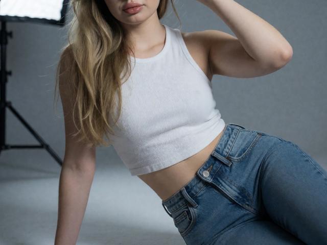Destinis with blonde hair, white crop top, and blue jeans, showing her slim waist and small tits, sitting with one arm raised, in a studio with soft lighting.