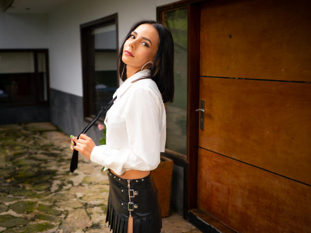 EvelinLuna with black hair, white shirt, black leather skirt with buckles, holding black strap, standing by wooden door, looking up with confident expression, medium skin, hoop earrings.