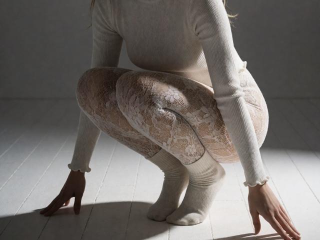 Destinis est accroupie, vêtue d'un haut blanc à manches longues en maille côtelée et de collants beiges à motifs de dentelle, avec des chaussettes blanches, et on voit ses fesses et ses mains sur un plancher en bois blanc..