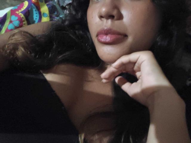 Paulette Delacroix, con i capelli ricci scuri, le labbra rosa lucide e un viso leggermente sudat, appoggia una mano vicino al mento, mostrando la spalla scoperta e la parte superiore del petto..